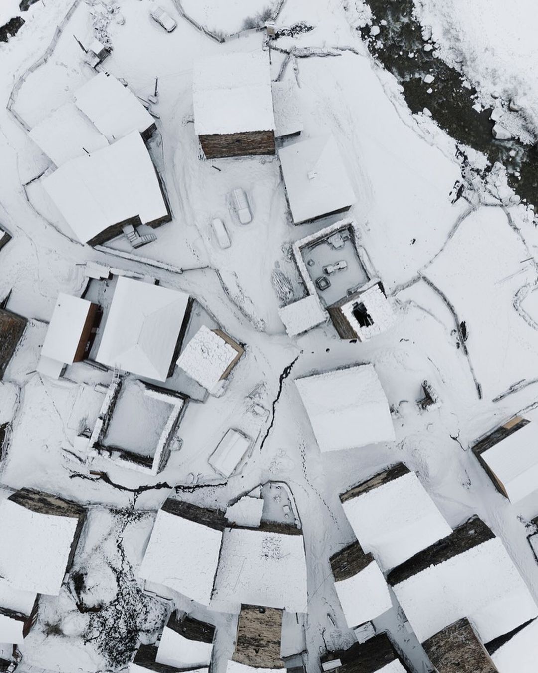 Aerial view of a snow-covered village with stone buildings and a winding stream, creating a serene winter scene in a rural environment.