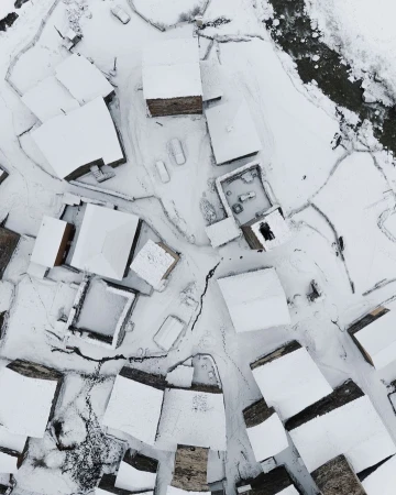 Aerial view of a snow-covered village with stone buildings and a winding stream, creating a serene winter scene in a rural environment.