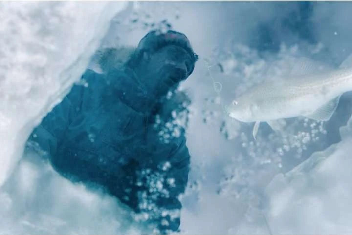 Underwater view of a fisherman in winter gear looking down through a hole in the ice at a fish swimming nearby in clear blue water.