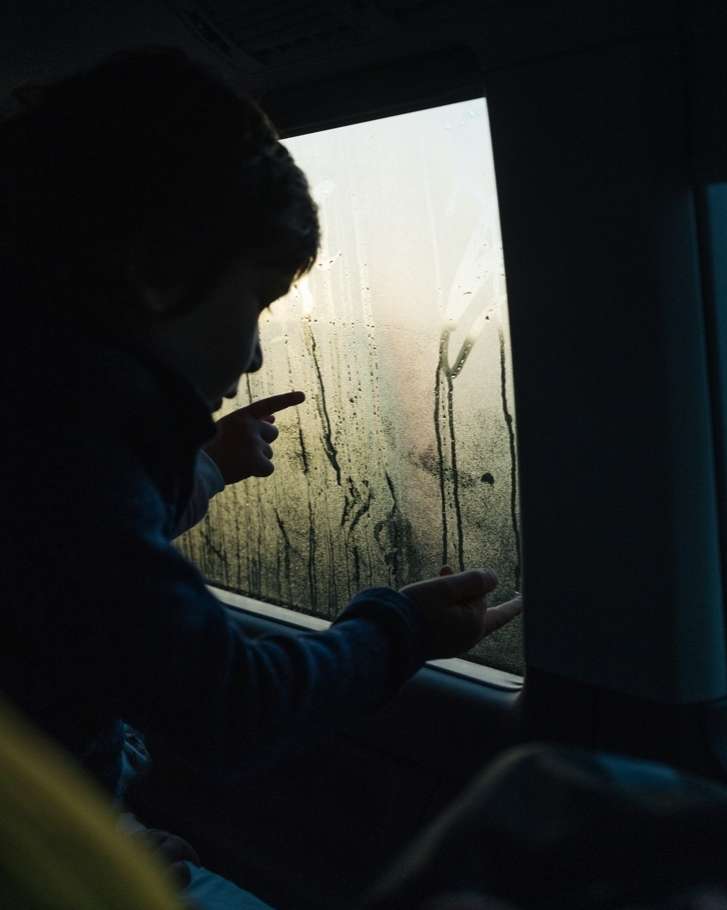 Child drawing on a fogged car window with fingers; streaks of light pass through the blurred background outside the glass.