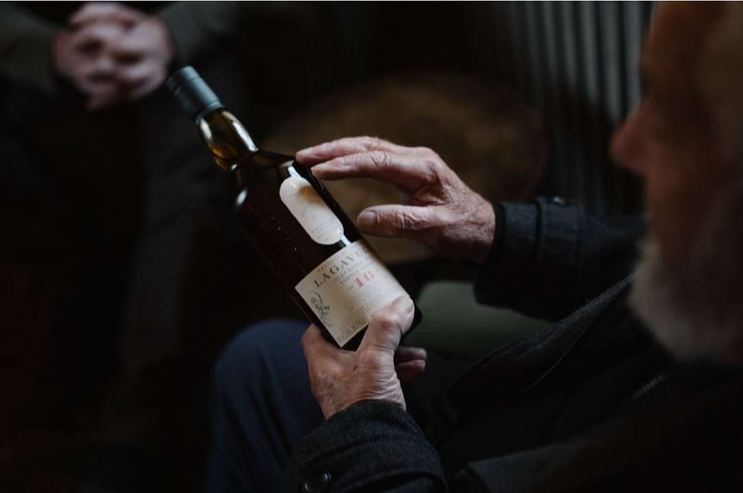 Close-up of an older man holding a bottle of Lagavulin 16 single malt Scotch whisky indoors, focused on the bottle and his hands.