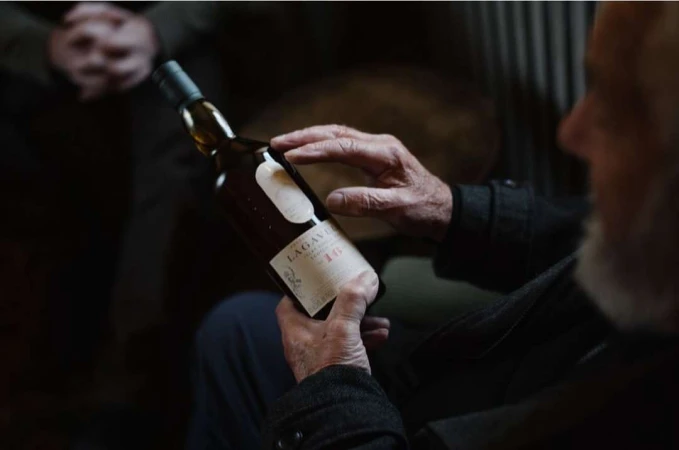 Close-up of an older man holding a bottle of Lagavulin 16 single malt Scotch whisky indoors, focused on the bottle and his hands.