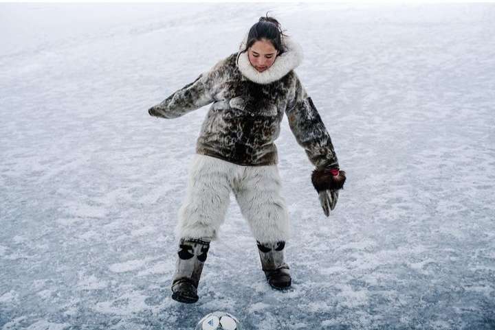 Young woman plays soccer on a frozen lake or field, wearing a fur coat, light fur pants and boots on a cloudy day in a winter landscape.