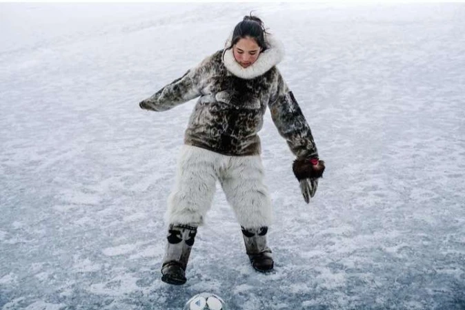 Young woman plays soccer on a frozen lake or field, wearing a fur coat, light fur pants and boots on a cloudy day in a winter landscape.