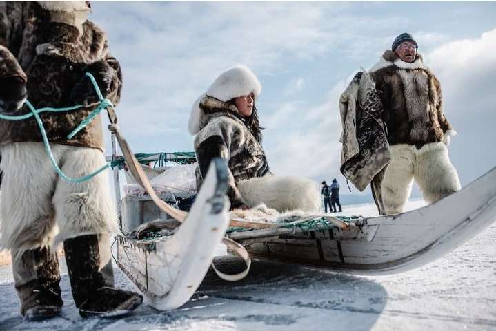 Three individuals wearing fur clothing ride on a sled in a snowy outdoor setting against a bright sky, evoking a cold environment.