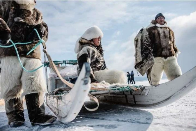 Three individuals wearing fur clothing ride on a sled in a snowy outdoor setting against a bright sky, evoking a cold environment.