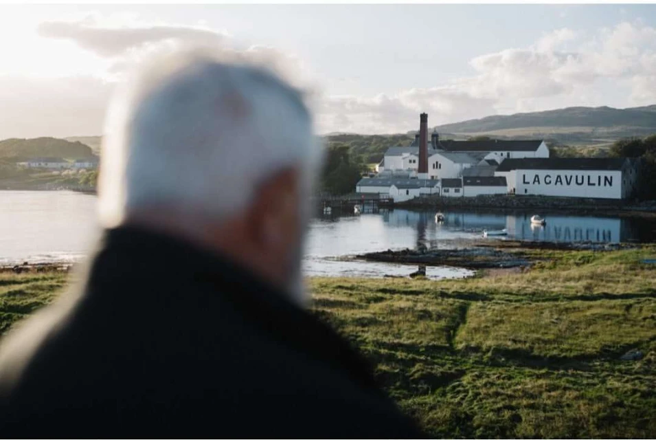 Distant view of the Lagavulin distillery in Scotland overlooking a body of water, with an adult wearing dark clothing in the foreground.