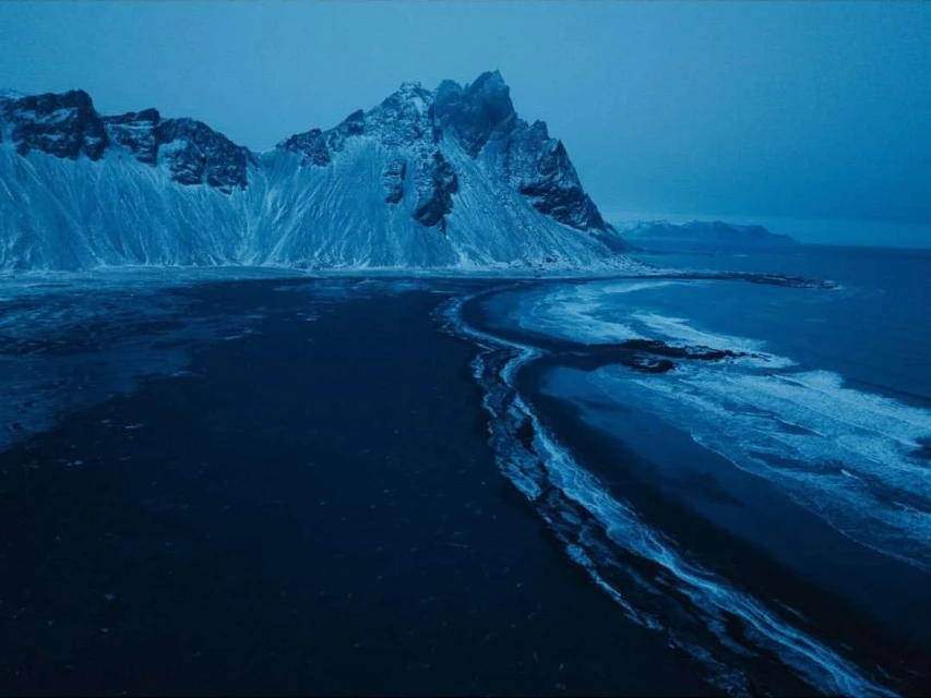 Dramatic aerial view of snow-covered Vestrahorn mountain range in Iceland meeting the dark ocean, shot in dim blue light.