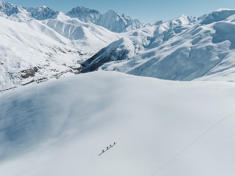 High-angle shot of four people walking on a snow-covered mountain landscape under a clear blue sky, featuring jagged snowy peaks.