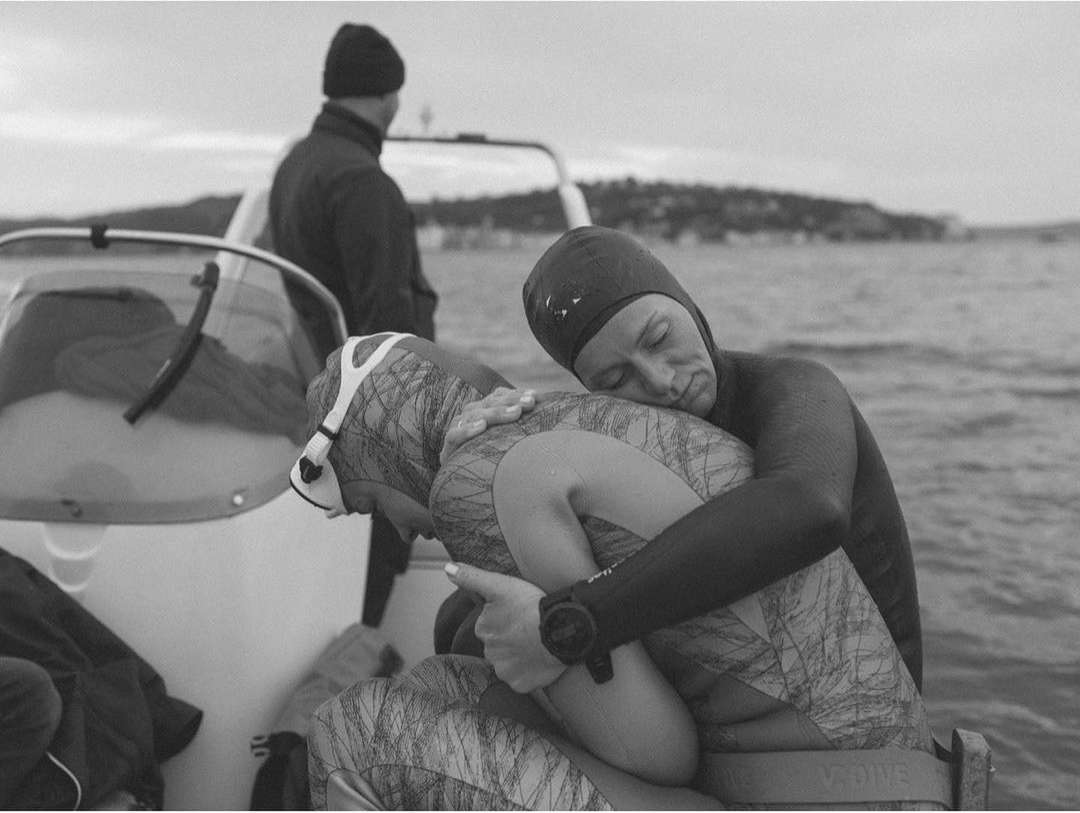 Two free divers in wetsuits on a boat embrace, with another person visible in the background against an ocean and sky backdrop.
