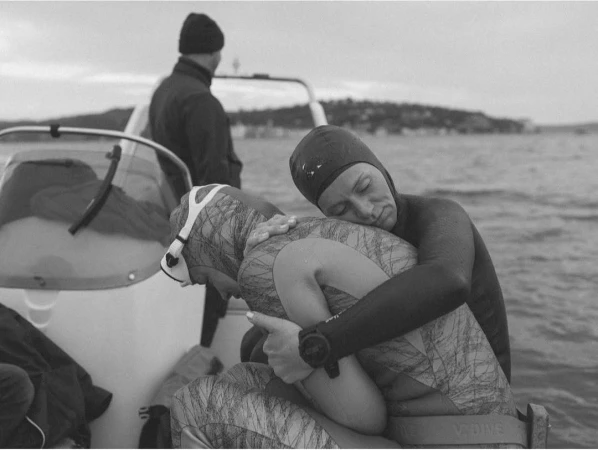 Two free divers in wetsuits on a boat embrace, with another person visible in the background against an ocean and sky backdrop.