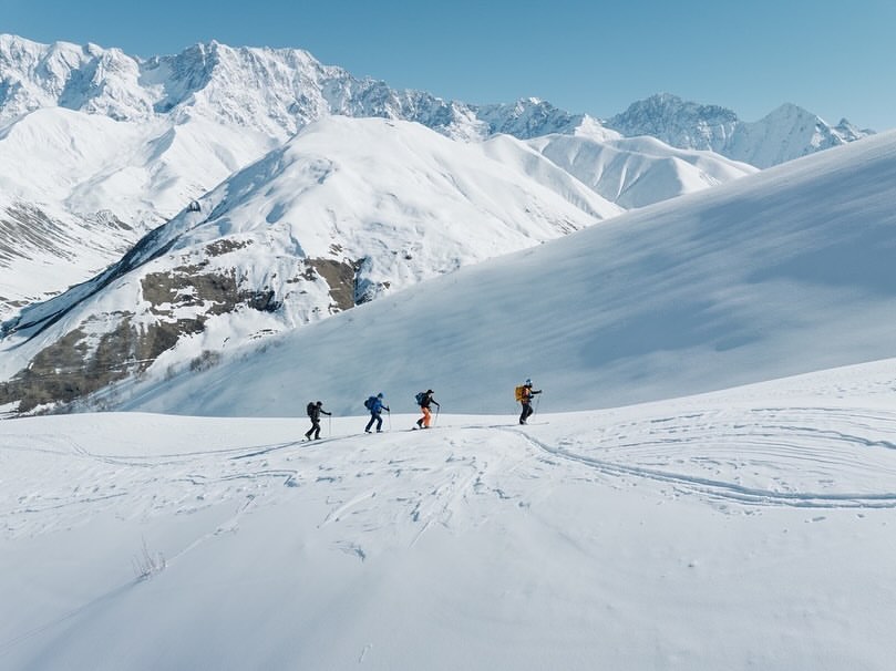 A group of four people skiing in a snowy mountain landscape under a clear sky, captured in a wide shot on a bright, sunny day.