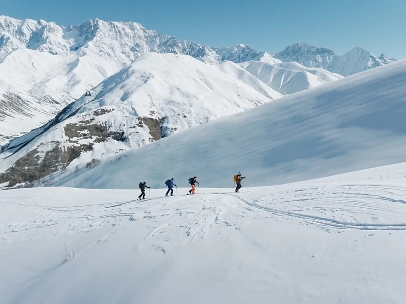 A group of four people skiing in a snowy mountain landscape under a clear sky, captured in a wide shot on a bright, sunny day.
