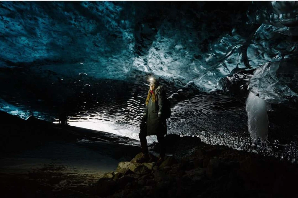 Adult female explorer stands atop rocky mound inside a glacial cave illuminated by a headlamp, with textured blue-ice walls and an icy floor.
