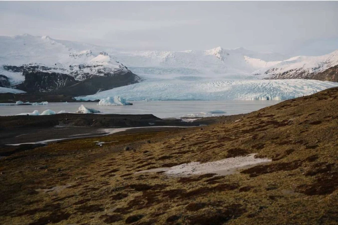 Landscape view of a glacial lake with icebergs under a cloudy sky, framed by a grassy hillside, presenting a cold, remote Arctic scenery.