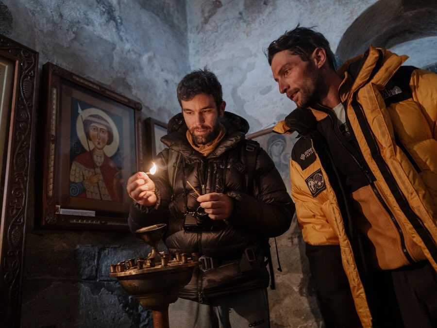 Two men inside a stone building, one lighting a candle; icons on the wall behind. The man on the right wears a yellow puffer jacket.