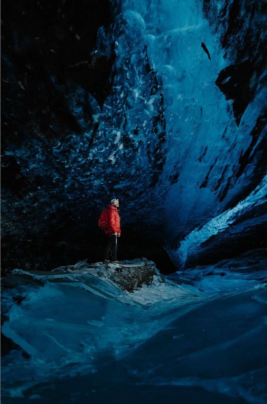Person in a red jacket stands inside a striking blue ice cave, gazing at the naturally sculpted ice formations and patterns.