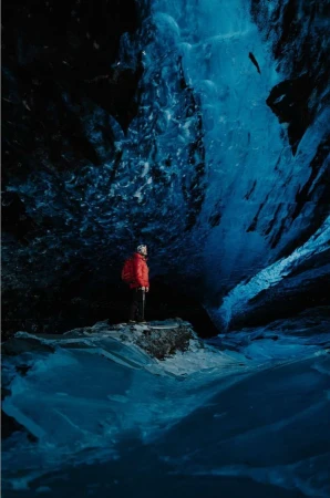 Person in a red jacket stands inside a striking blue ice cave, gazing at the naturally sculpted ice formations and patterns.