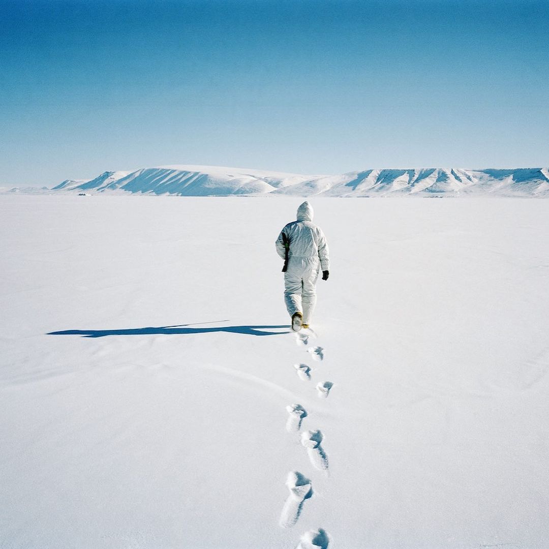 Person in a white hazmat suit walks across a snowy plain towards distant mountains under a clear blue sky, leaving footprints.