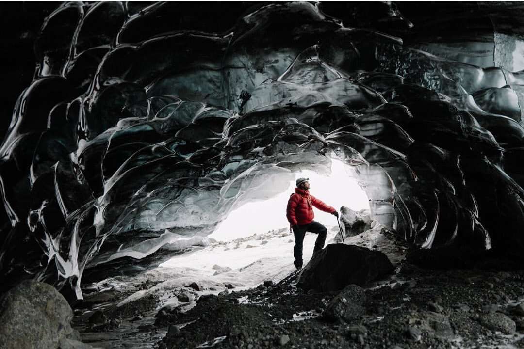 Man in red jacket exploring a glacier cave with ice formations, wearing a headlamp, standing with an ice axe on a rocky surface.