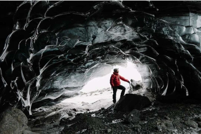 Man in red jacket exploring a glacier cave with ice formations, wearing a headlamp, standing with an ice axe on a rocky surface.