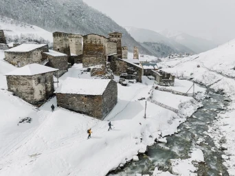 Aerial view of skiers on a snow-covered path next to a river with ancient stone towers in a mountain village