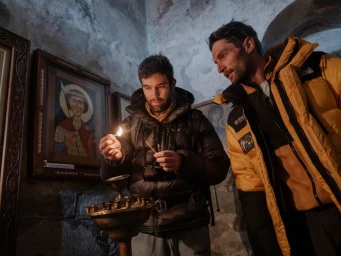 Two men standing inside an ancient stone church, one lighting a match over a candle holder next to a framed religious icon.