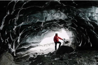 An explorer in a bright red jacket standing inside a dark, icy cave mouth, looking out toward a bright snowy landscape.