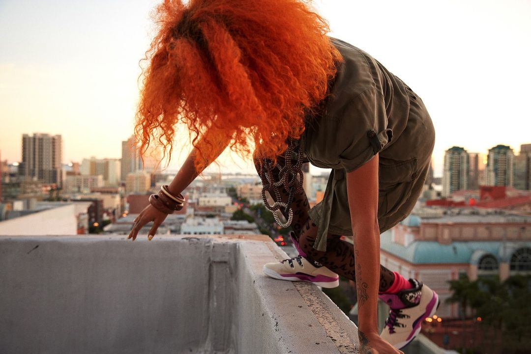 Woman with orange hair crouches on a rooftop, wearing patterned leggings, with a cityscape visible at sunset in the background.