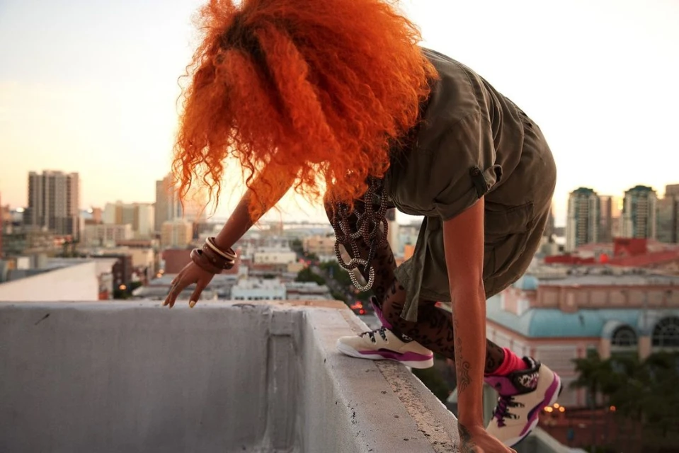 Woman with orange hair crouches on a rooftop, wearing patterned leggings, with a cityscape visible at sunset in the background.