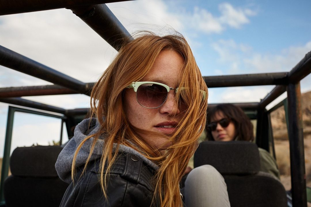 Woman with red hair and sunglasses rides in the back of an off-road vehicle with another passenger in a desert landscape under a cloudy sky.