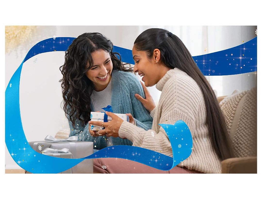 Two smiling women are indoors during daytime, one giving a wrapped gift to the other. They are sat on a couch with blue graphic design.