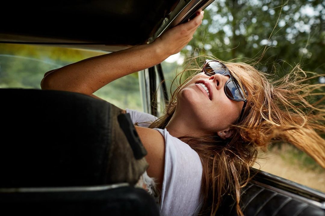 Woman with sunglasses leans out car window, wind in her hair, traveling on a sunny day, trees visible in the background.