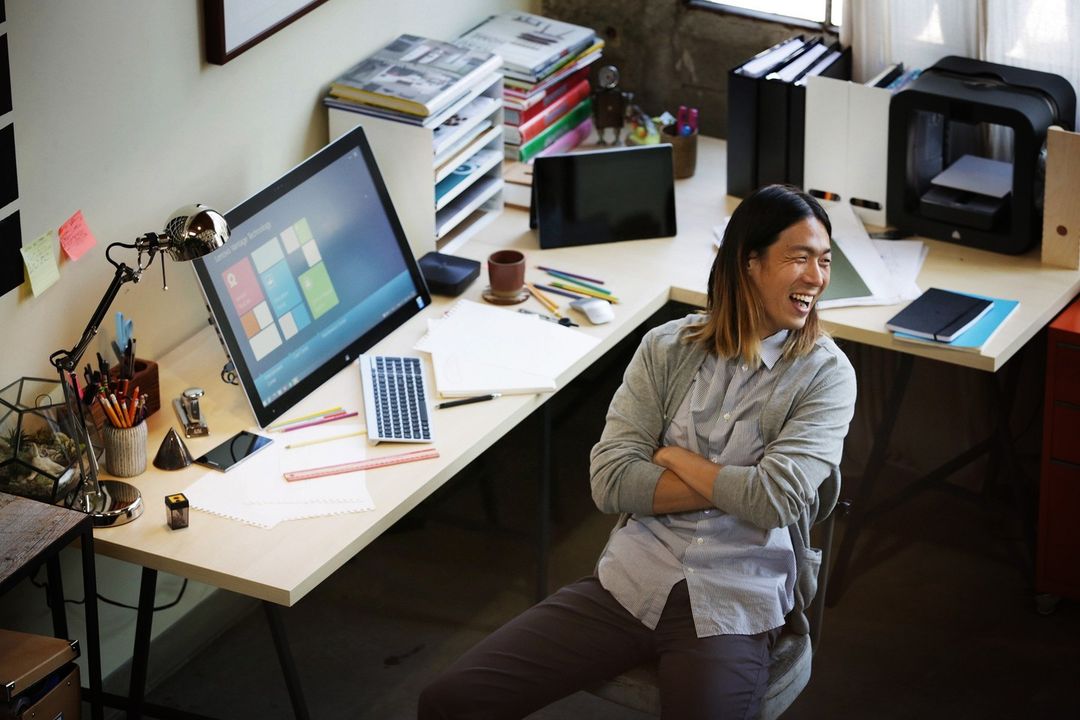 Smiling man sits at a bright office desk with computer, office supplies, books, and a 3D printer creating a modern workplace scene.