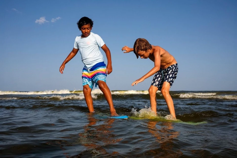 Two boys enjoy skimboarding on a sunny day at the beach. The boys are standing on their boards in the water with the ocean in the background.