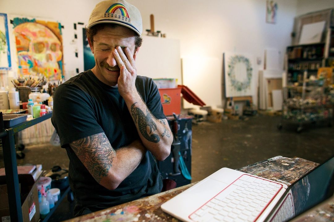Artist in studio laughing with hand over face near paint-covered desk, laptop. Background art, painting supplies fill the bright space.