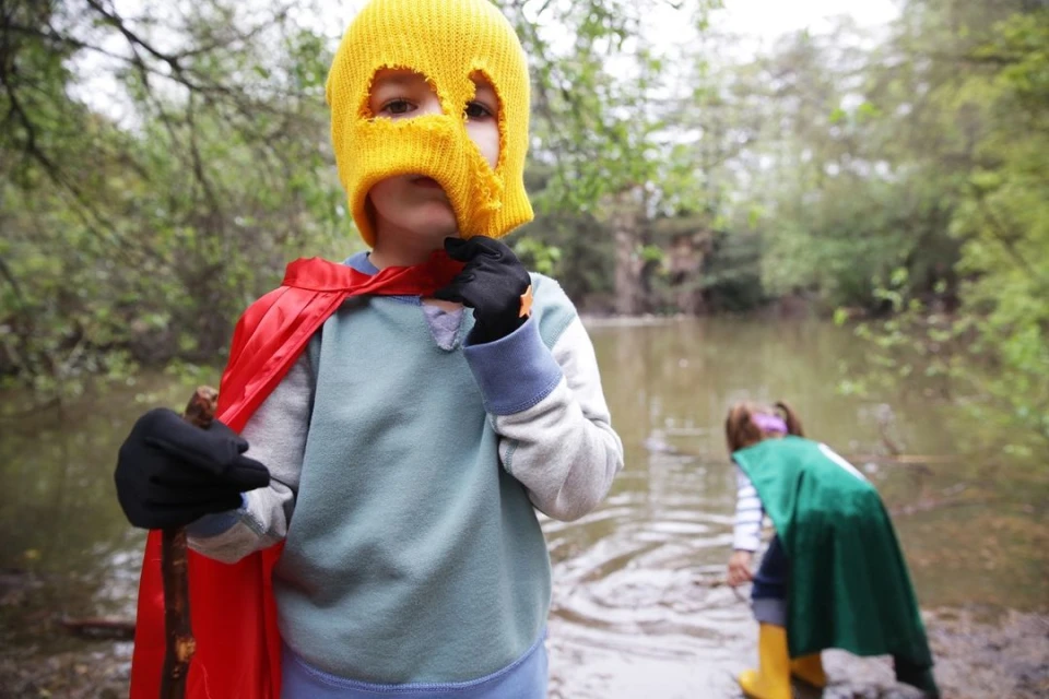 Child in yellow balaclava and red cape stands near water with another child in green, trees in the background, outdoor setting.