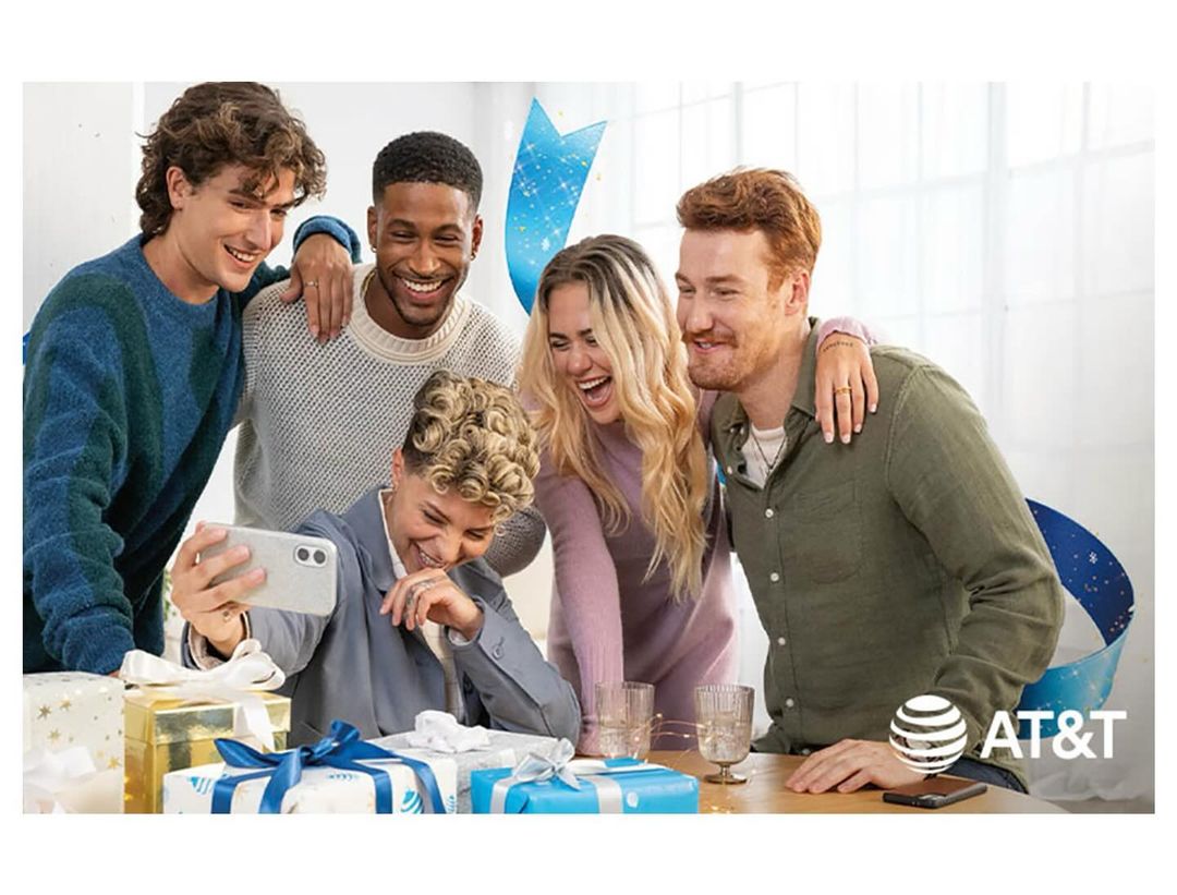 Group of five friends gather around a table with gift boxes, laughing and taking a selfie together. An AT&T logo is visible.