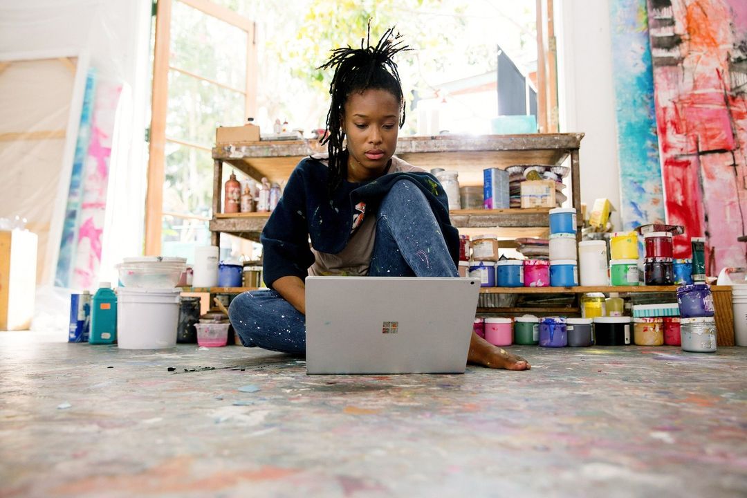 An artist with dreadlocks sits cross-legged on a paint-splattered floor, working on a laptop in a brightly lit, creative studio space.