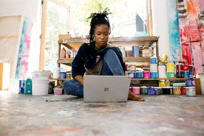 An artist with dreadlocks sits cross-legged on a paint-splattered floor, working on a laptop in a brightly lit, creative studio space.