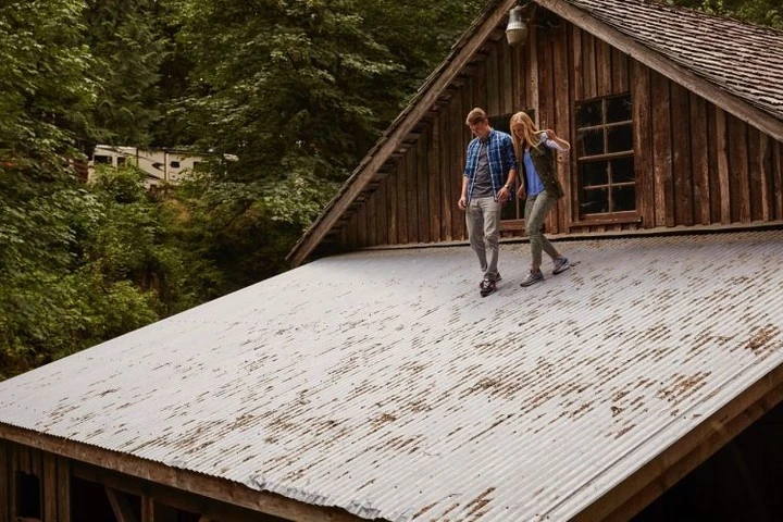 A fair-haired couple carefully walk on the corrugated metal roof of a wood cabin with a forest and camper in the background.