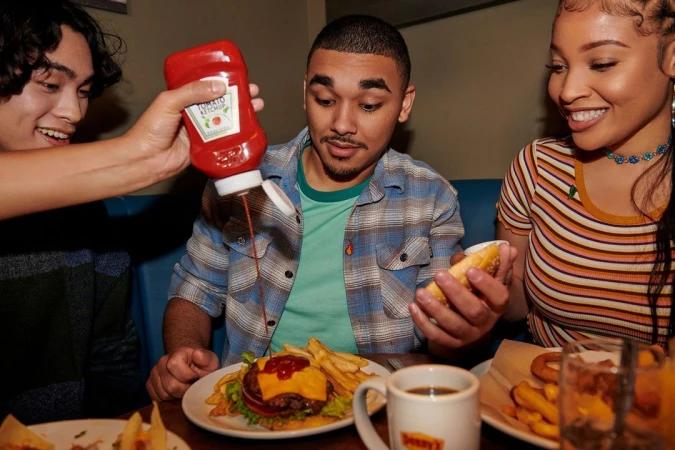 Three people at a diner table with a burger and fries, one person pouring ketchup from a Heinz bottle, all smiling and having a meal.