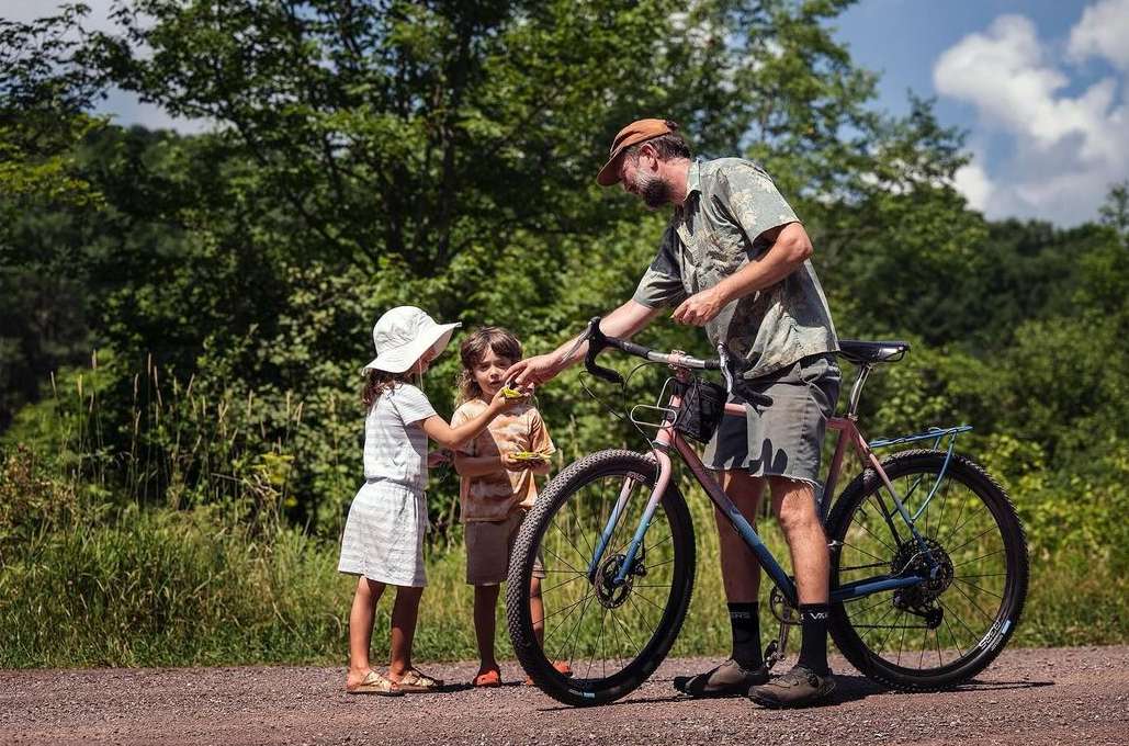A father shares something with his two young daughters, standing alongside a bicycle on a dirt path in a green, tree-filled environment.