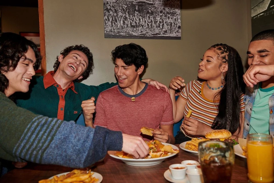 Five young adults laughing and eating together at a casual restaurant with burgers, french fries, and nachos on a wood-grain table.