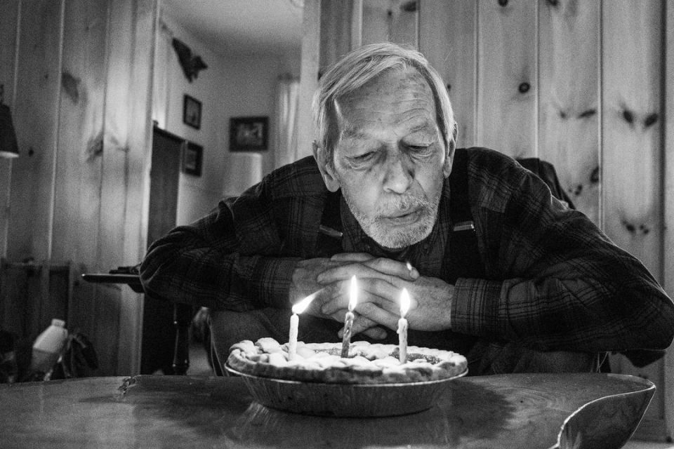 Elderly man blowing out candles on a pie in a monochrome indoor shot. He is wearing a flannel shirt with suspenders.