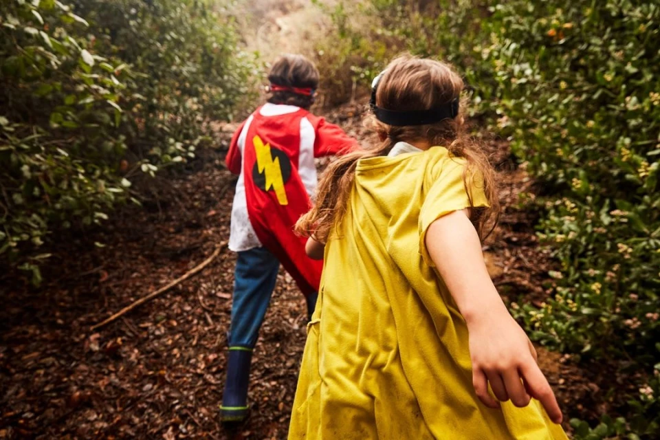 Two children wearing superhero capes navigate a woodland trail, the bright colors contrasting with the natural setting.