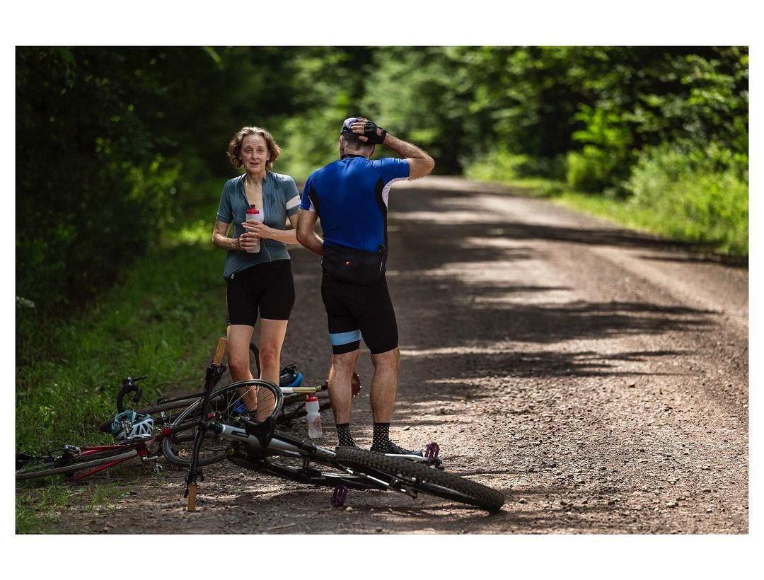Two cyclists take a break on a gravel road in a forest, with one holding a water bottle and a bicycle lying on the ground.