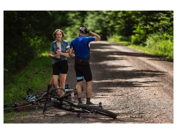 Two cyclists take a break on a gravel road in a forest, with one holding a water bottle and a bicycle lying on the ground.