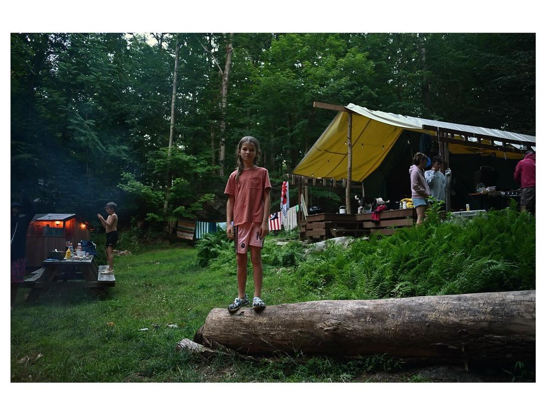 A young girl stands on a log in an outdoor setting near a tent and lush greenery, capturing a casual and candid moment in nature.
