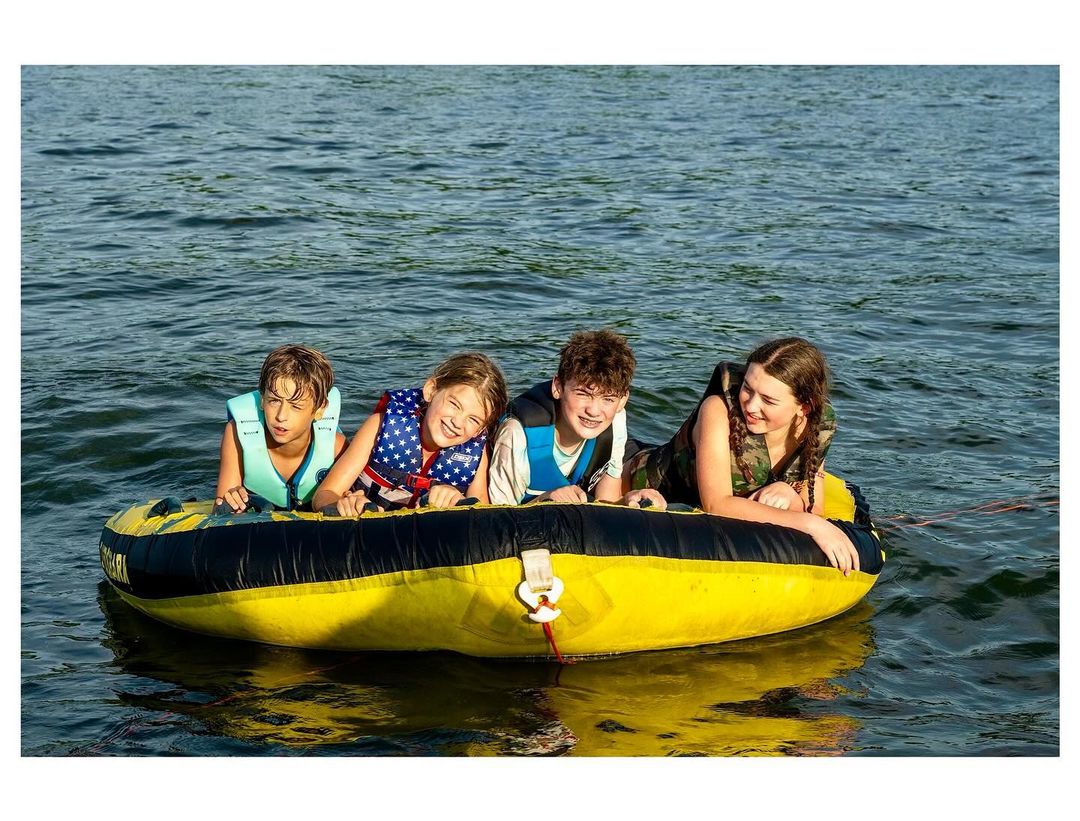 Four children in life vests are riding in a large yellow inflatable tube on a lake during the daytime; a watersport adventure.